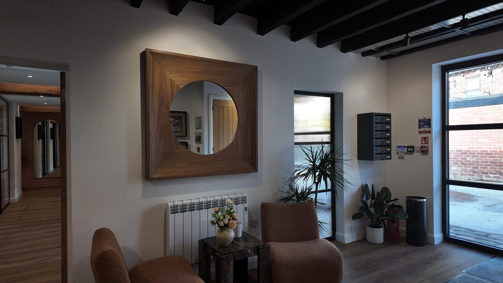 The breakout corner by the entrance: two armchairs beneath a round walnut-framed mirror, with plants and the post boxes on the wall.