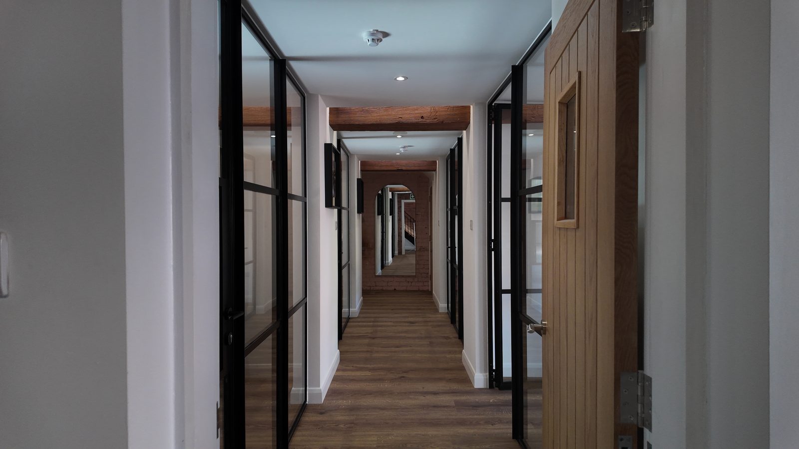 The corridor: black-framed glazed partitions either side of an oak floor, an original timber beam overhead and a mirror on brick at the end.