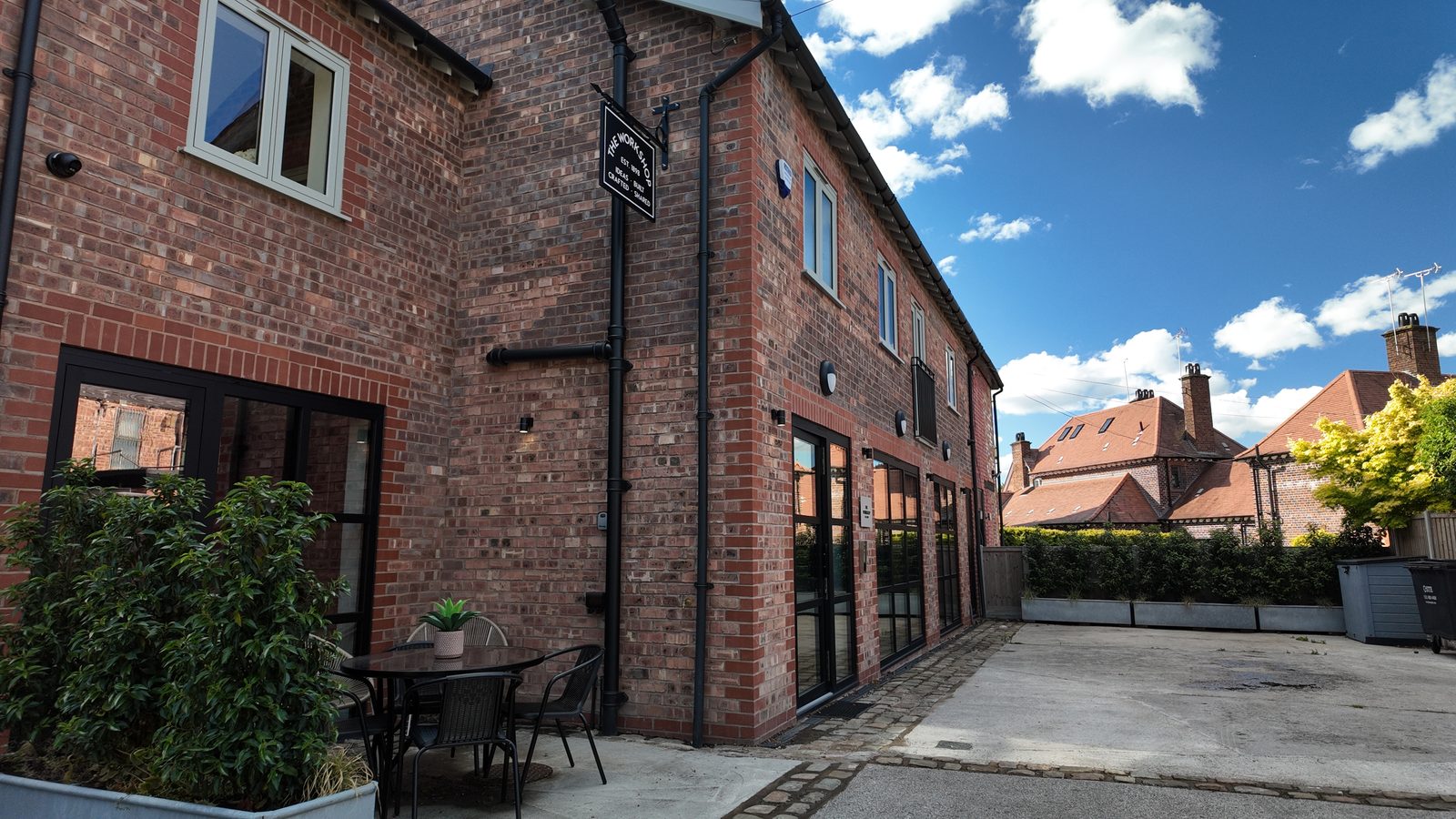 The courtyard: a bistro table and chairs against the brick corner of the building, with planters along the drive and blue sky above.
