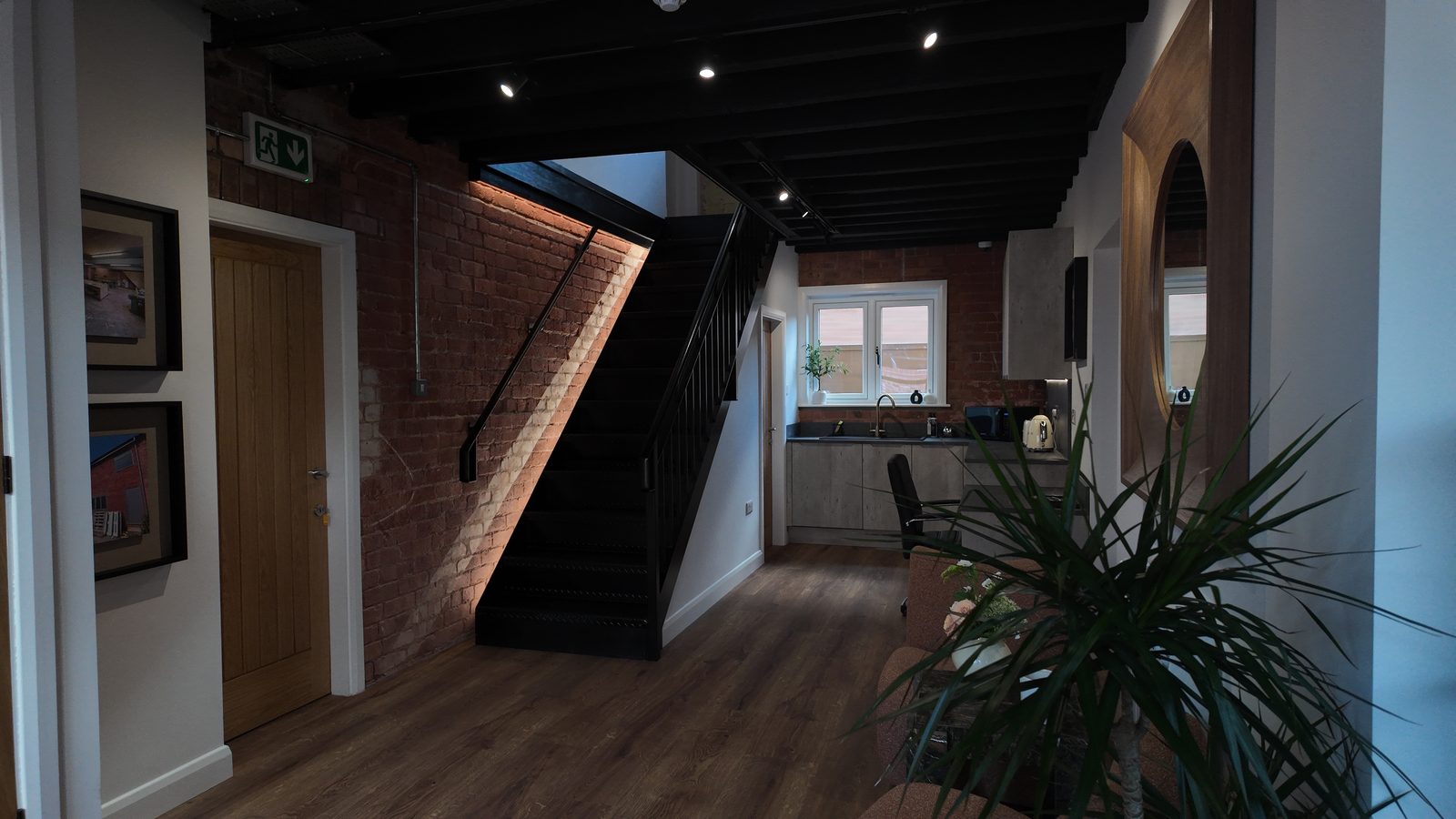 The entrance hall: a black steel staircase against lit exposed brick, with an oak floor and the shared kitchen beyond.