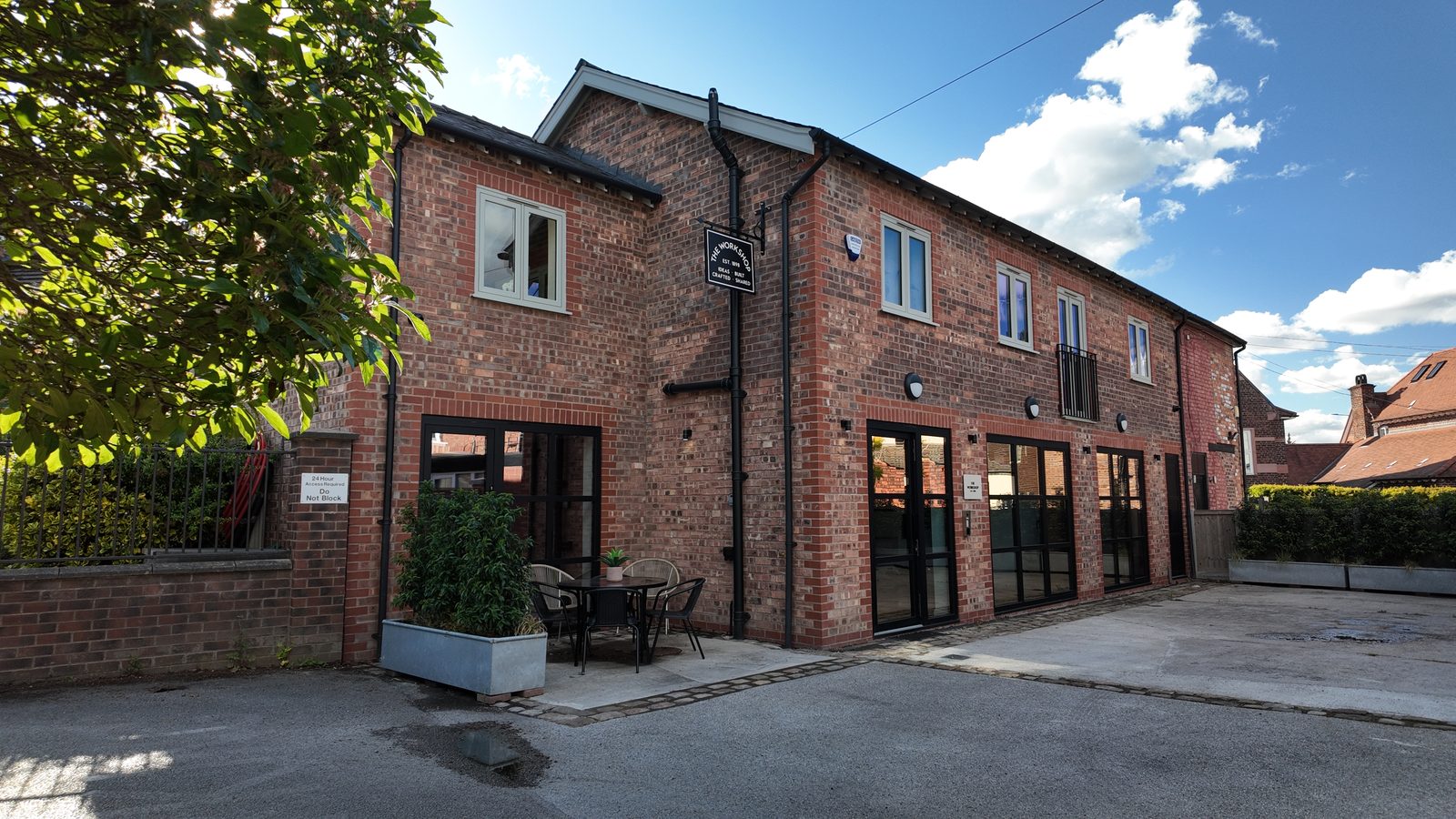 The brick front elevation of The Workshop in evening light, with black Crittall-style glazing, the hanging sign and a bistro table in the courtyard.