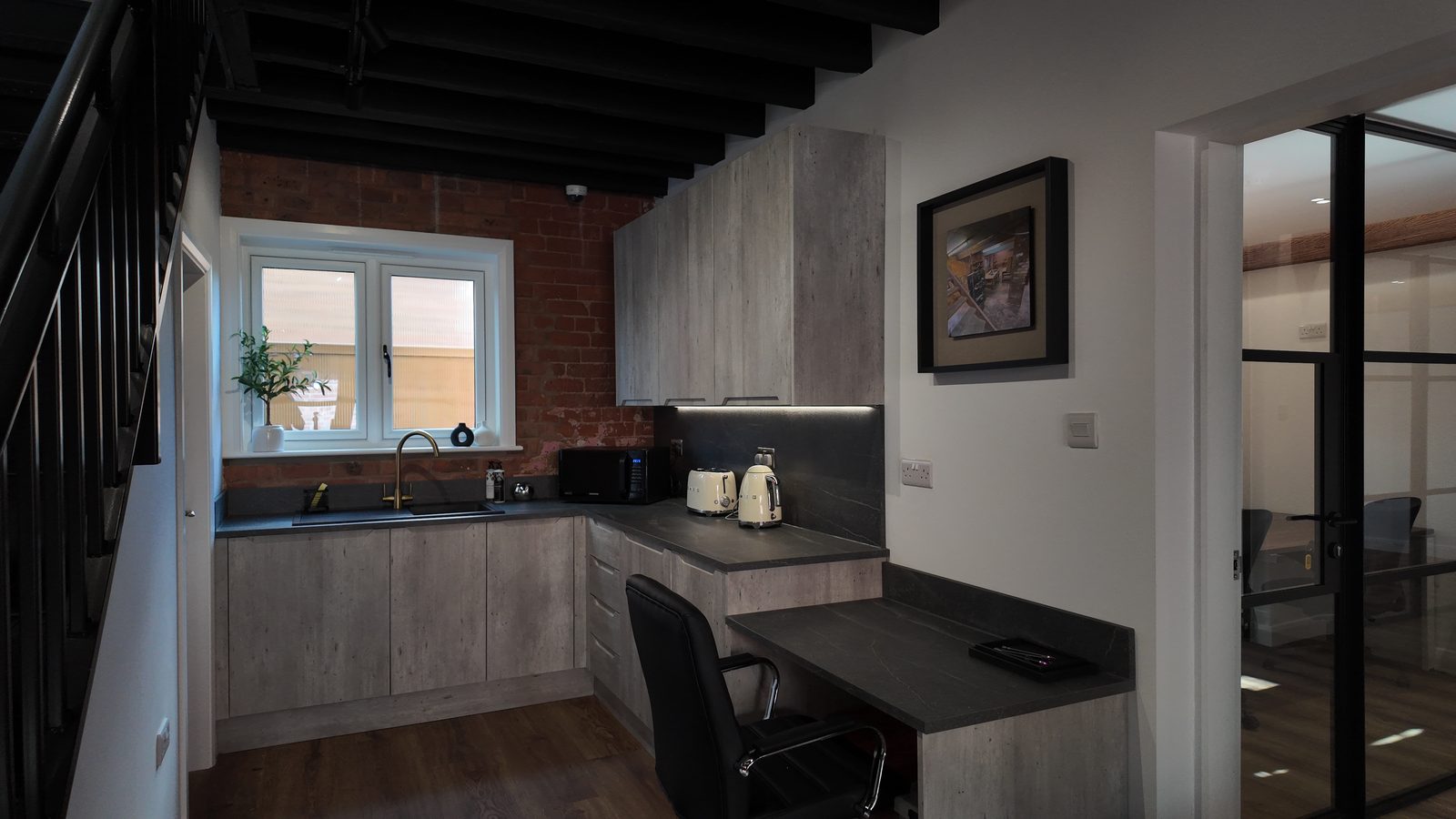 The shared kitchen: black-painted beams, an exposed brick splashback, concrete-effect worktops and a window over the sink.