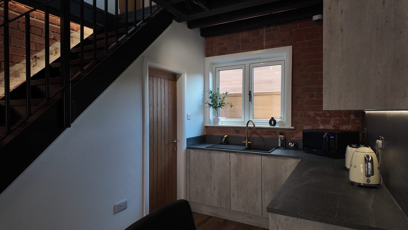 The kitchen from the foot of the stairs: a brass tap on a stone-effect worktop, brick splashback and a cream kettle.