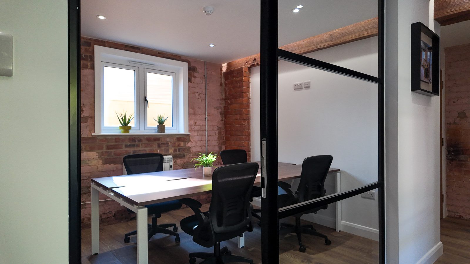 Office 6, seen from its glazed door: two desks face to face with four chairs, plants on the windowsill and exposed brick.