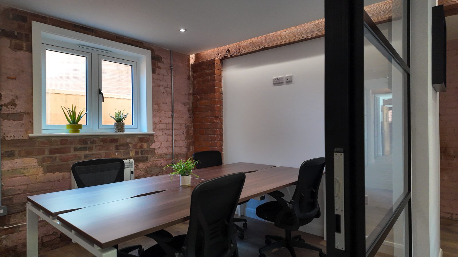 Office 7: bench desks with chairs between exposed brick walls, a window with plants on the sill and a black-framed glazed door.