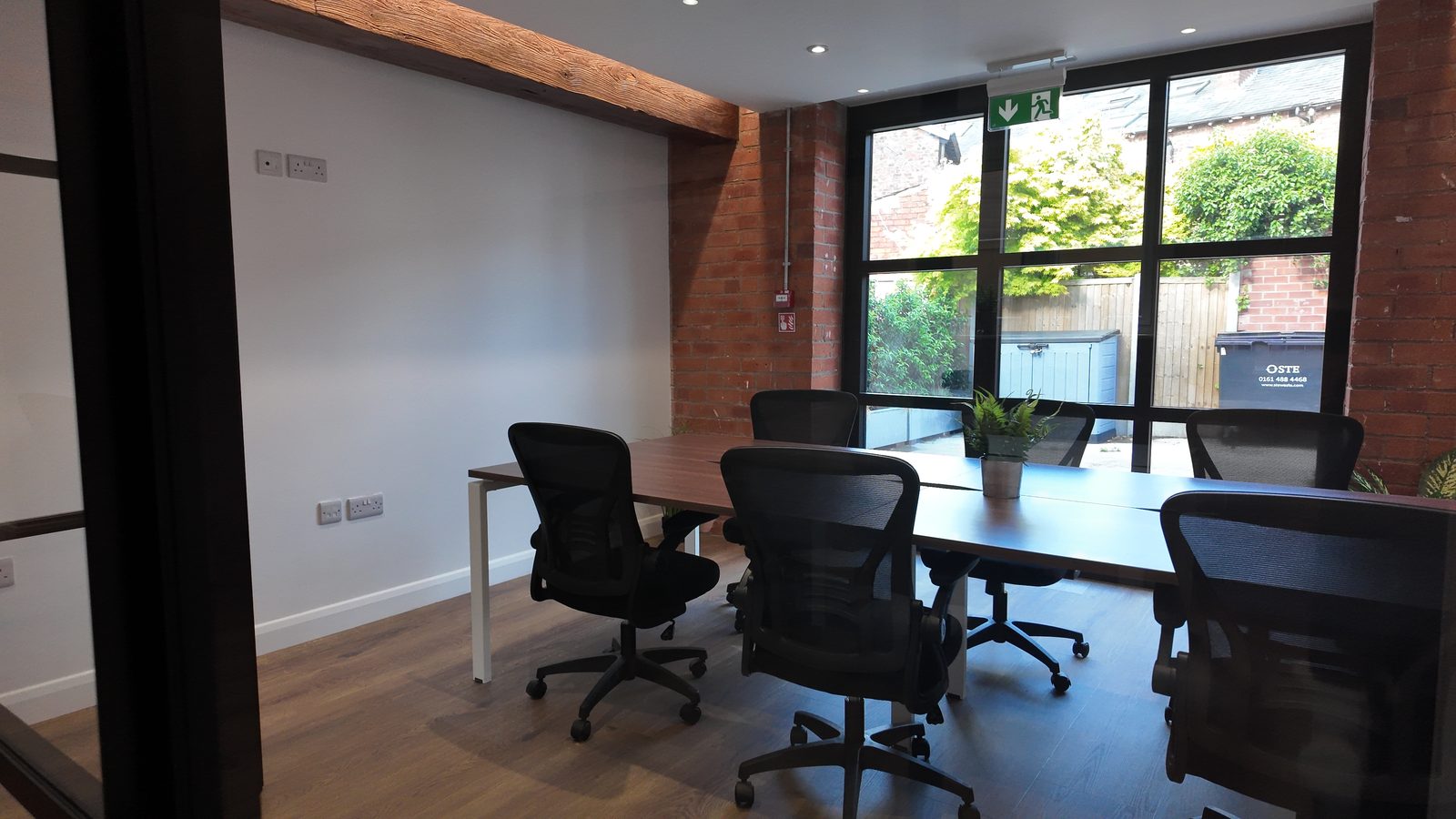 Office 9: a shared desk island with six chairs, an exposed brick pillar and full-height glazing onto the courtyard planting.