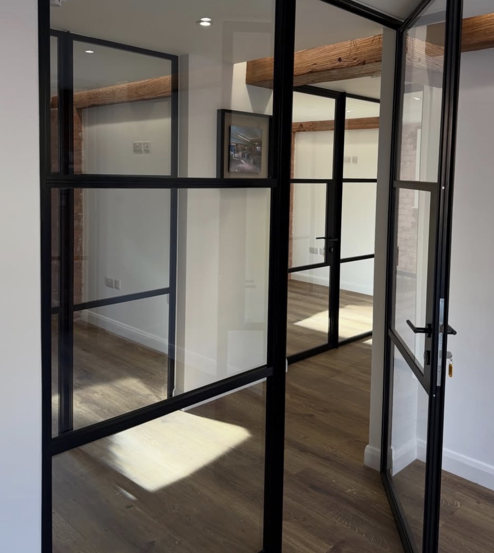 Black-framed glazed partitions and an open glazed door beneath a reclaimed timber beam, with sunlight pooling on the oak floor.