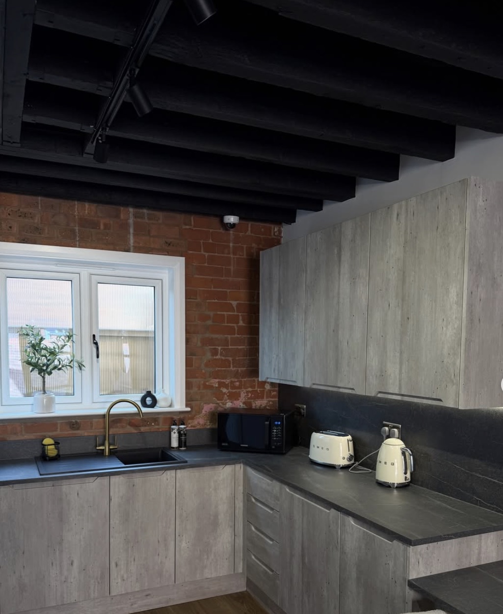 The shared kitchen: black-painted beams, exposed brick, concrete-effect units and a brass tap.