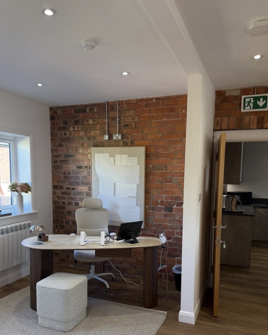 An office room with a brick feature wall, an oval walnut desk and a view through to the shared kitchen.