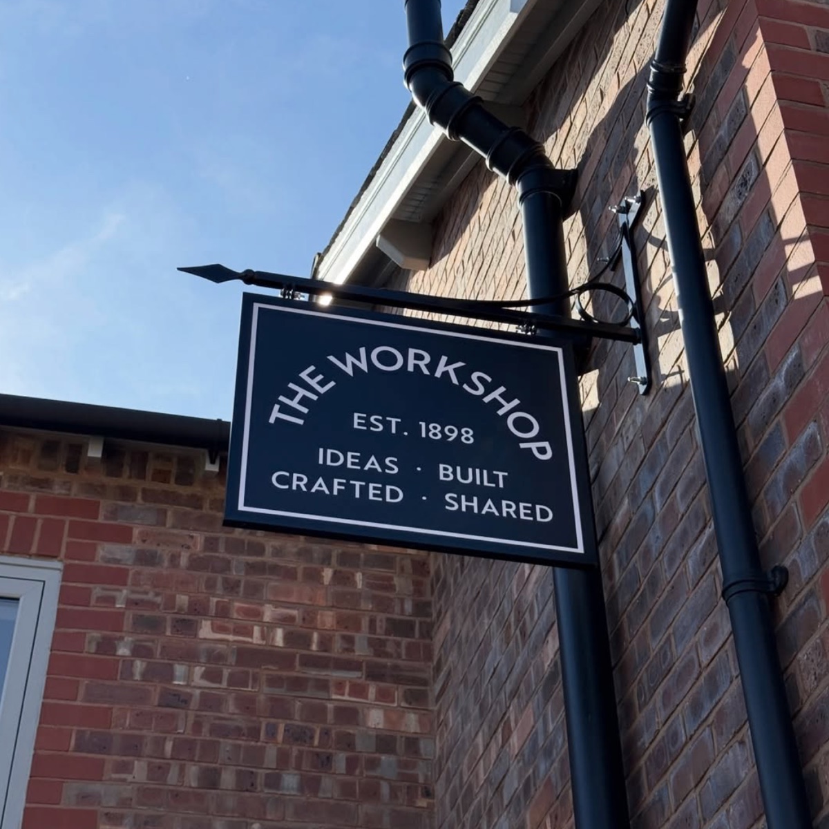 The navy hanging sign against a blue sky: The Workshop, Est. 1898, Ideas Built, Crafted Shared.