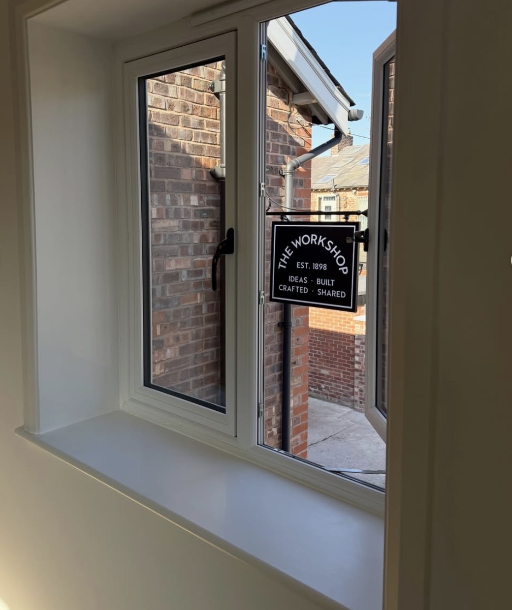 The navy hanging sign seen through an open window, with the brick outbuildings beyond.