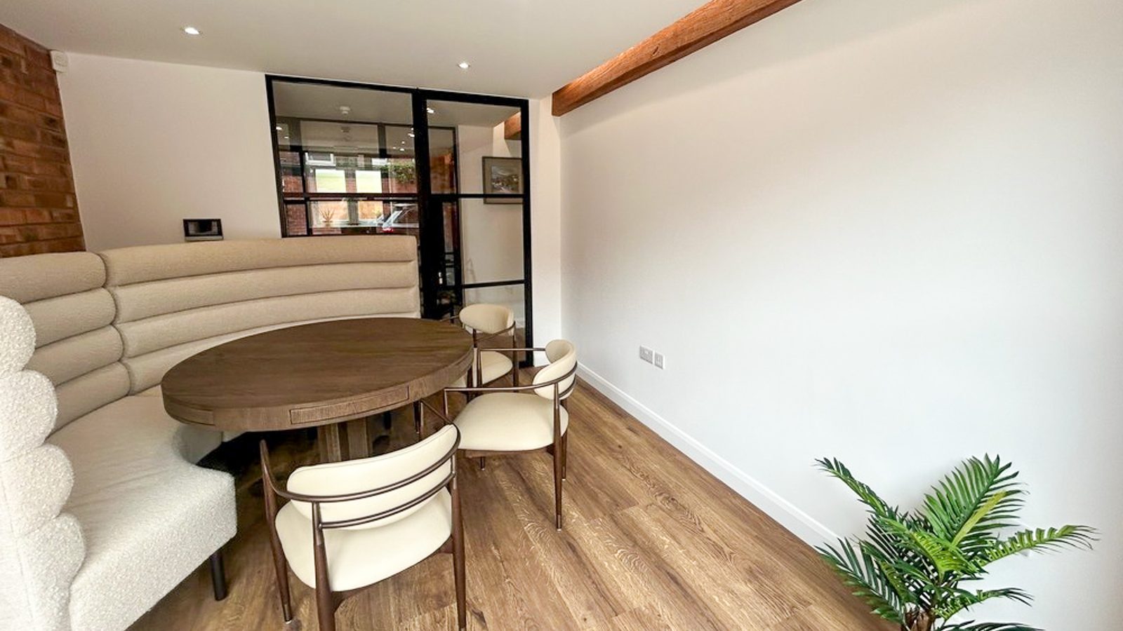 The Meeting Room: a curved cream banquette and a round oak table with cream chairs, a glazed black-framed partition behind, exposed brick and a timber beam.